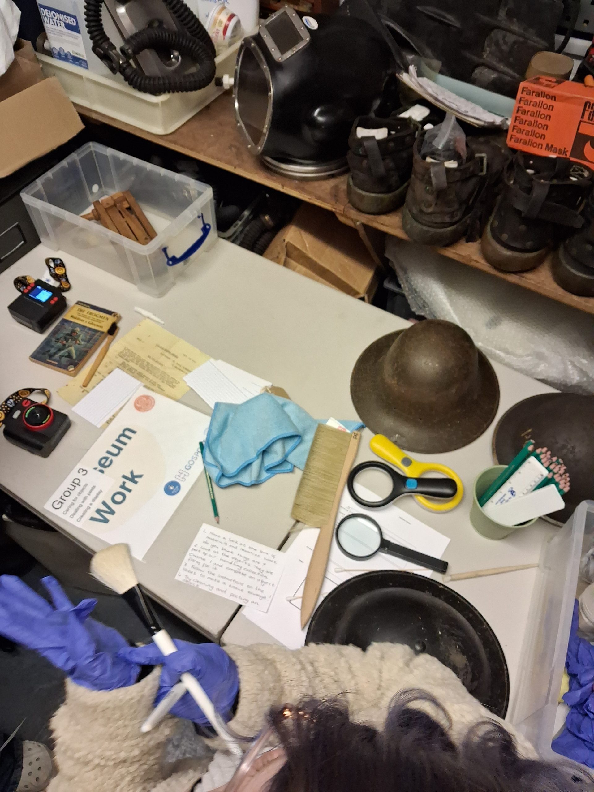 A table with stell helmets, magnifying glasses, cameras, brushes and papers with weighted diving boots in the background